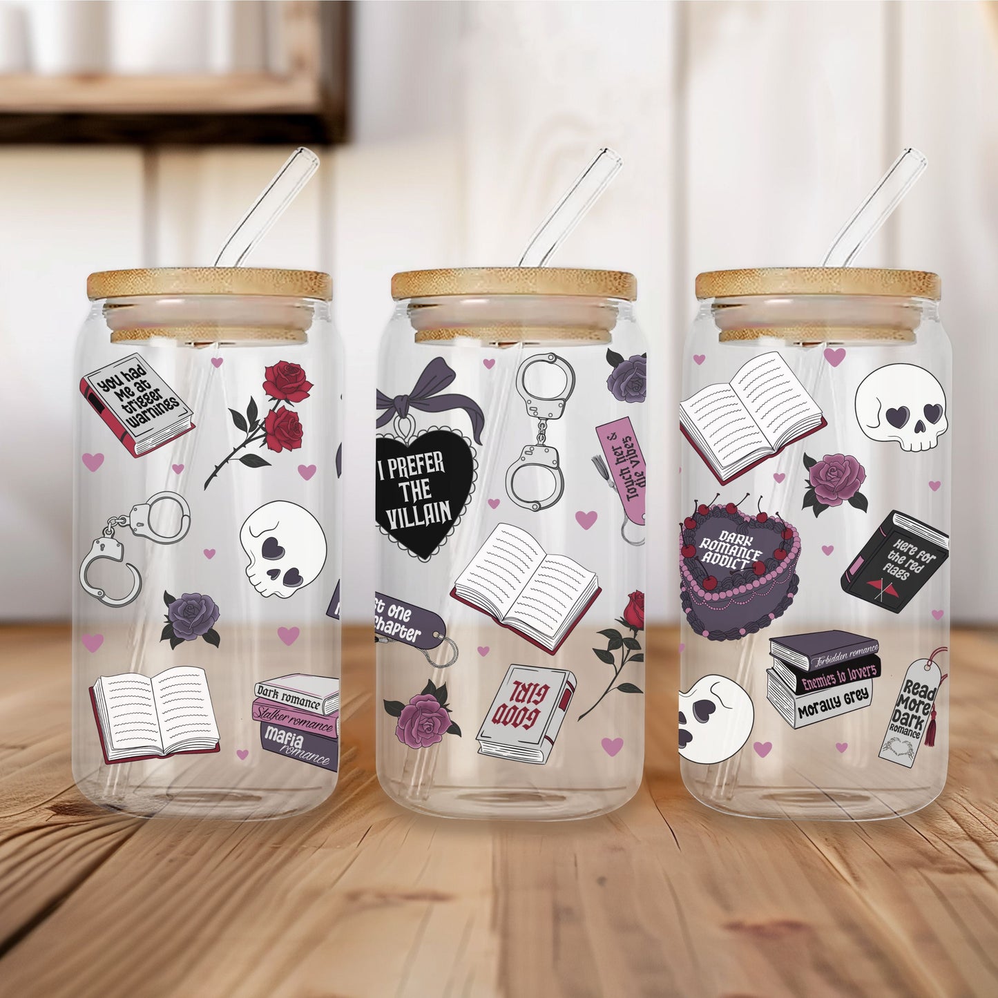 Three clear glass mason jar tumblers with bamboo lids and glass straws, decorated with dark romance themed DTF stickers including skulls, open books, roses, and hearts, displayed on a wooden table.